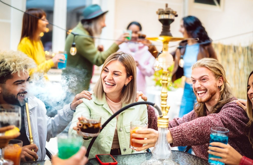 A group of young adults sit around a table outdoors, laughing and clinking colorful drinks. A large hookah sits in the center of the table, and people in the background are also socializing. The scene is lively and cheerful, with warm lighting and a relaxed atmosphere.