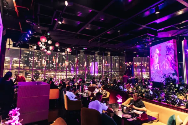 A vibrant, modern restaurant interior with purple and pink neon lighting, large windows overlooking a city skyline at night, and diners seated at stylish tables and booths. Hanging lights and contemporary artwork add to the lively, upscale atmosphere