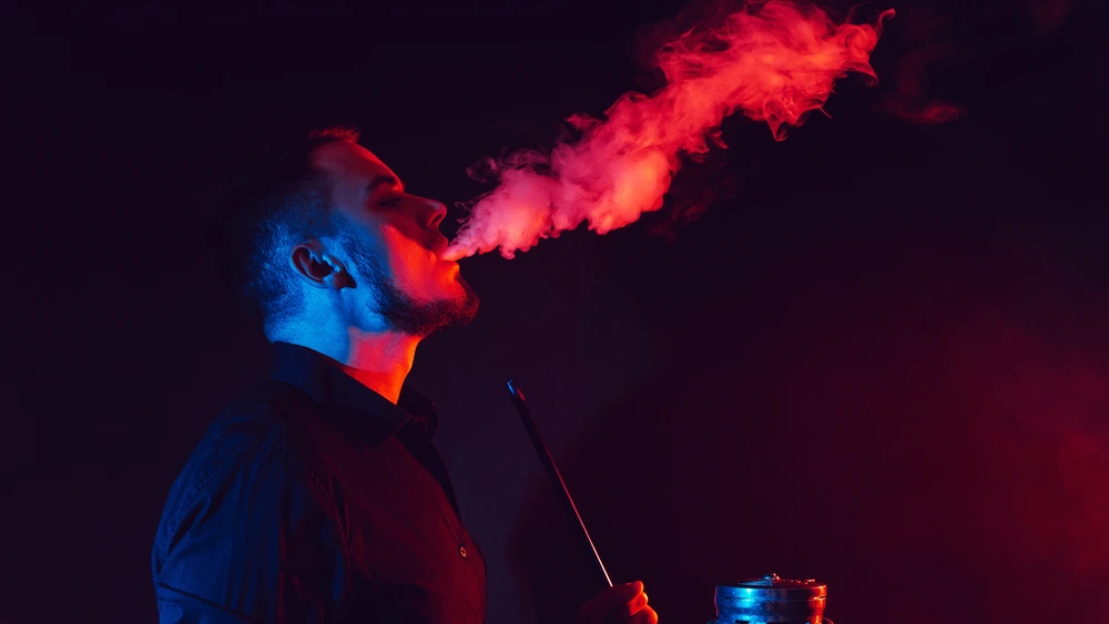 Man enjoying shisha, exhaling vibrant red smoke in a dark, artistic setting that reflects a luxury shisha experience in Dubai.