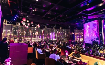 A vibrant, modern restaurant interior with purple and pink neon lighting, large windows overlooking a city skyline at night, and diners seated at stylish tables and booths. Hanging lights and contemporary artwork add to the lively, upscale atmosphere